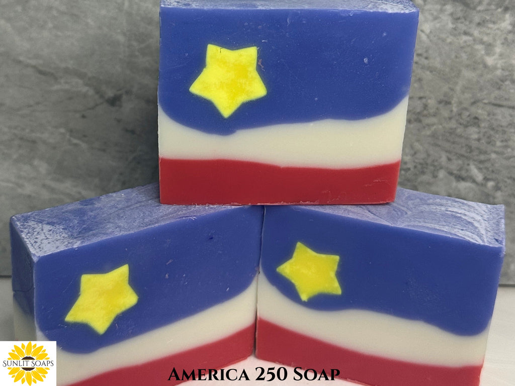 Three bars of soap with a patriotic design featuring red, white, and blue colors and yellow stars on a gray surface. Sunlit Soaps logo in the bottom left corner