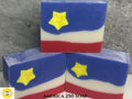 Three bars of soap with a patriotic design featuring red, white, and blue colors and yellow stars on a gray surface. Sunlit Soaps logo in the bottom left corner