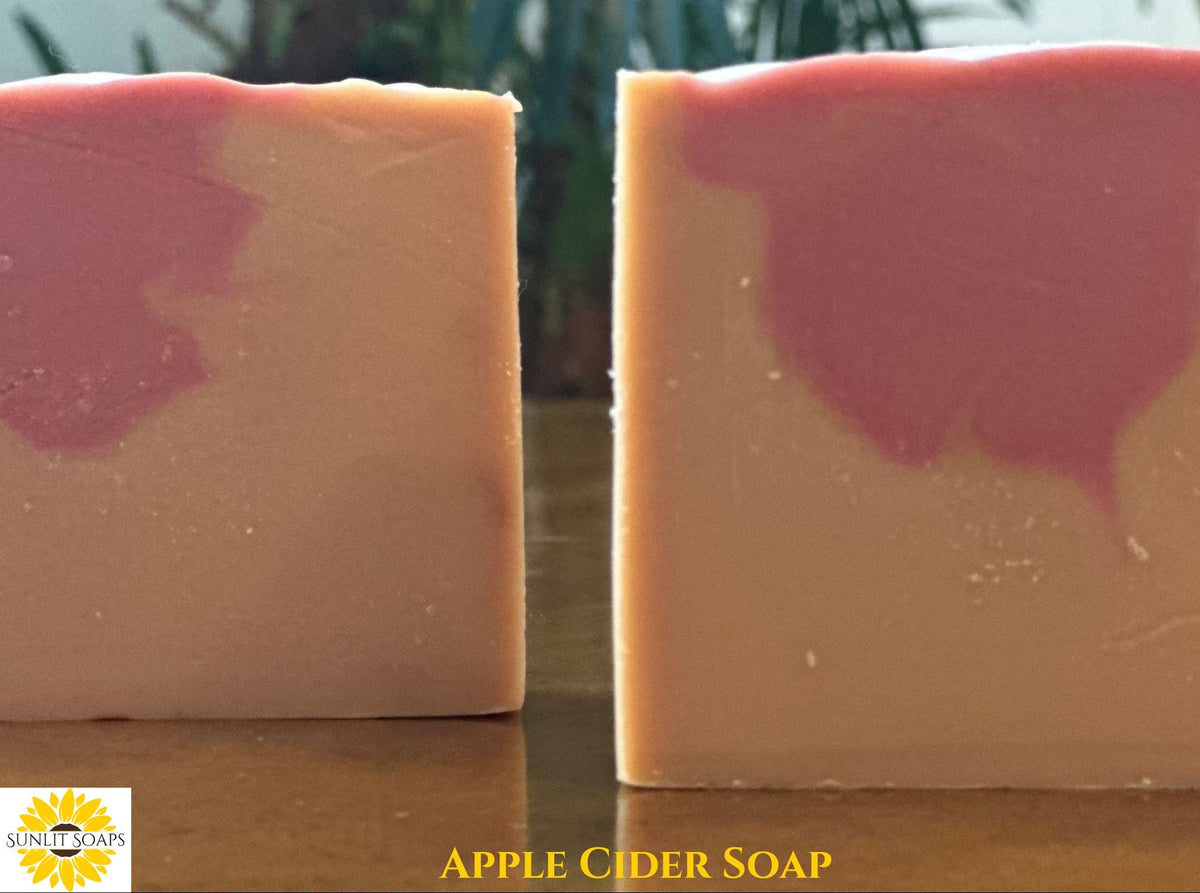 Two rectangular bars of apple cider soap standing upright on a wooden surface, with a pink swirl pattern on a warm peach-beige base. The left bar is partially cropped. The words "Apple Cider Soap" and the Sunlit Soaps logo is on the bottom edge.