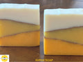 Two rectangular bars of layered Aspen Soap on a dark wooden surface. The soap features three horizontal color bands: a white top layer, a pale yellow middle layer, and a vibrant orange-yellow bottom layer, with the Sunlit Soaps logo in the bottom left corner