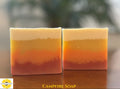 Two rectangular bars of layered handmade soap with horizontal bands of cream, yellow, orange, and reddish-brown, displayed on a wooden surface with a blurred outdoor background. The text 'CAMPFIRE SOAP' is visible at the bottom with the Sunlit Soaps logo in the left corner.