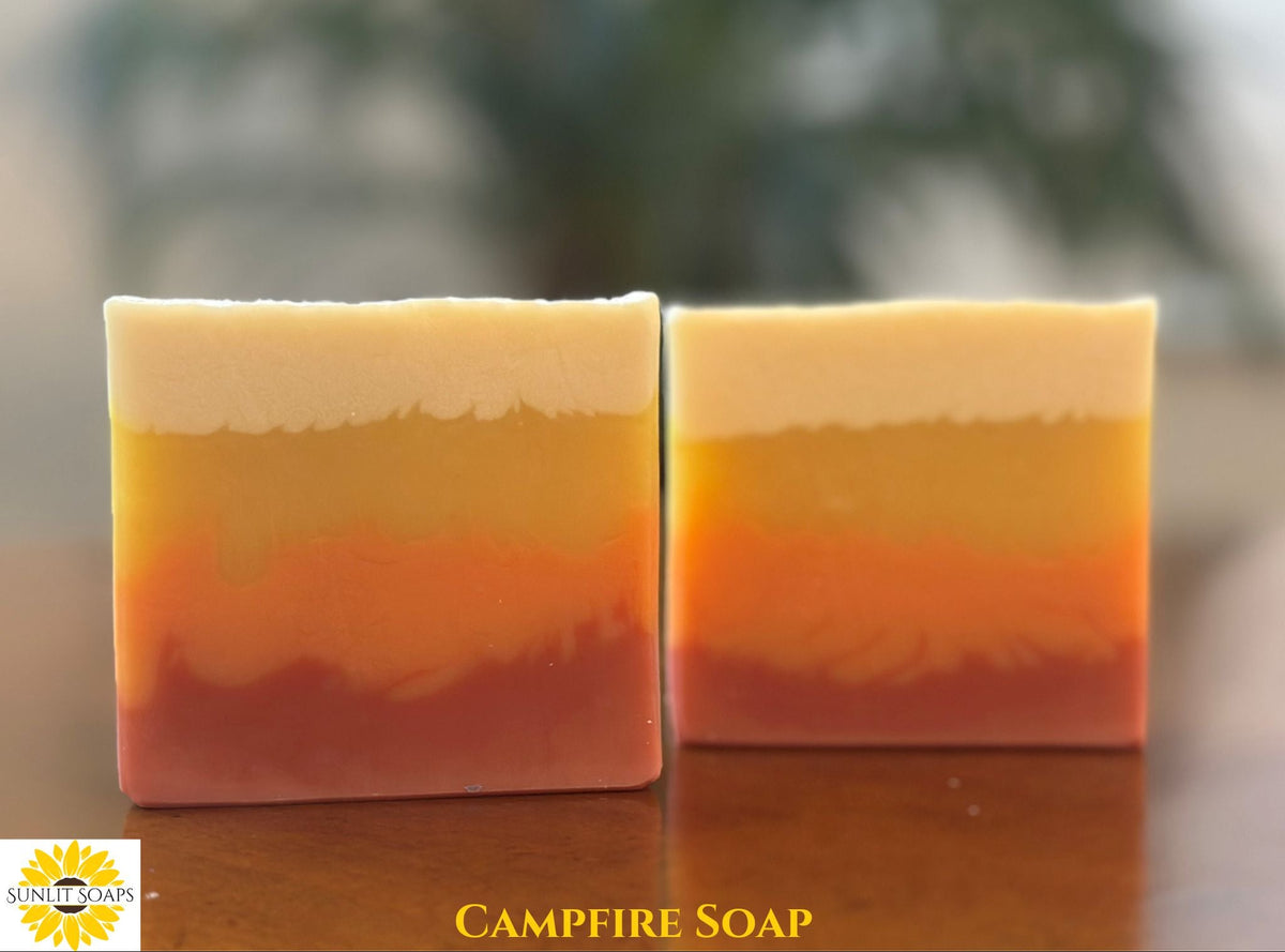 Two rectangular bars of layered handmade soap with horizontal bands of cream, yellow, orange, and reddish-brown, displayed on a wooden surface with a blurred outdoor background. The text 'CAMPFIRE SOAP' is visible at the bottom with the Sunlit Soaps logo in the left corner.