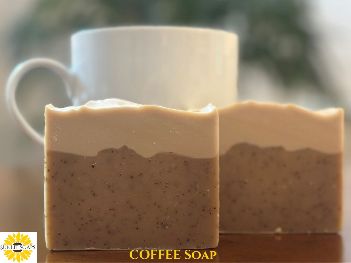 Two rectangular bars of handmade coffee soap in a layered beige and brown speckled design, displayed on a wooden surface with a white ceramic mug in the blurred background. A 'Sunlit Soaps' logo is visible in the bottom-left corner, and 'COFFEE SOAP' is written in yellow text at the bottom.