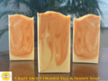 Three handmade soap bars with a swirled orange and cream pattern, displayed on a wooden surface with a blurred background of green leaves. A yellow logo for 'Sunlit Soaps' and the text 'CRAZY ABOUT ORANGE TALL & SKINNY SOAP' are visible at the bottom.