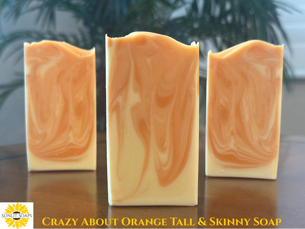 Three handmade soap bars with a swirled orange and cream pattern, displayed on a wooden surface with a blurred background of green leaves. A yellow logo for 'Sunlit Soaps' and the text 'CRAZY ABOUT ORANGE TALL & SKINNY SOAP' are visible at the bottom.