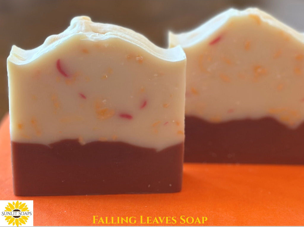 Two bars of handmade soap labeled 'FALLING LEAVES SOAP' by Sunlit Soaps, featuring a cream-colored top layer with small orange and red flecks and a dark brown bottom layer, set against a bright orange background.