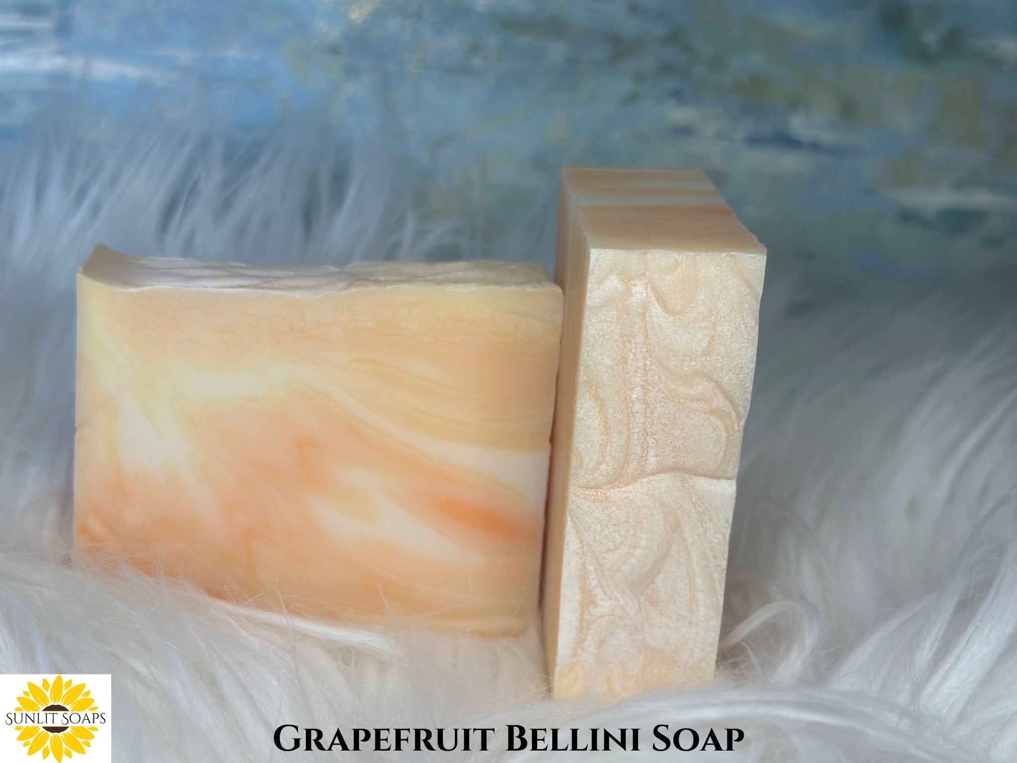 Two bars of Grapefruit Bellini soap on a textured white surface with a blurred blue background with Sunlit Soaps branding.