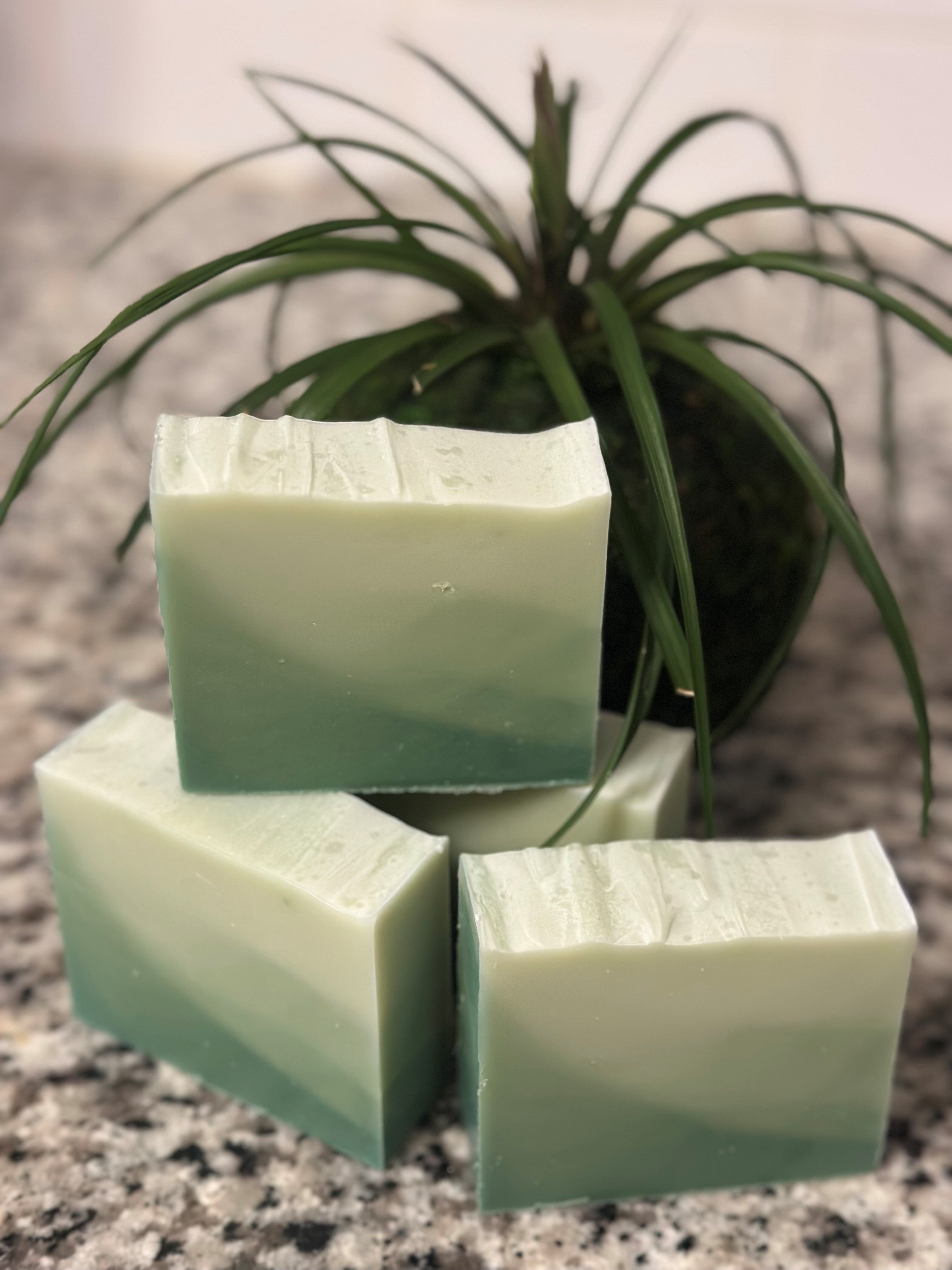 Three green ombre soap bars stacked on a speckled surface with a plant in the background. Branded title Sunlit Soaps.