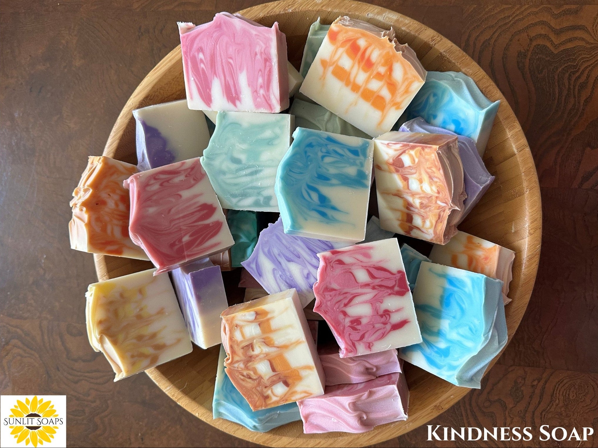 Assorted Kindness colorful soap bars in a wooden bowl on a wooden surface, with 'Sunlight Soaps' branding.