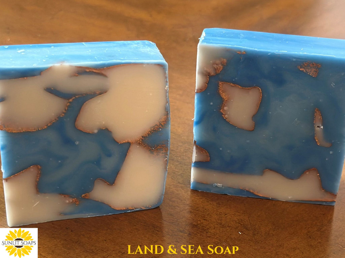 Two rectangular bars of handmade soap with a blue and cream swirl pattern and copper-colored edges, displayed on a wooden surface. A logo for 'Sunlit Soaps' is visible in the bottom-left corner, and the text 'LAND & SEA SOAP' is written at the bottom.