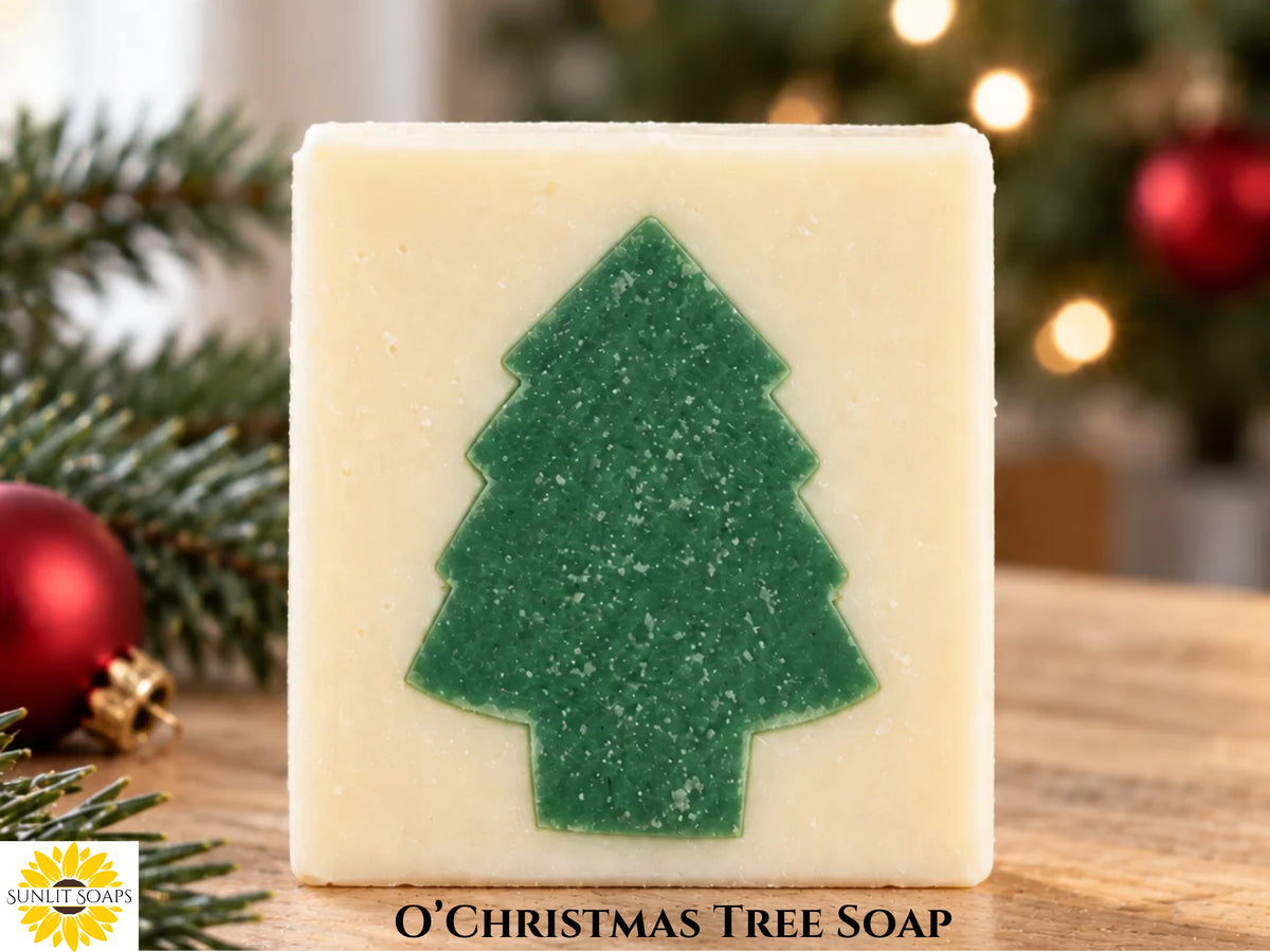 Ivory colored bar of soap with a green tree shape in the center on a wooden table. Christmas decorations and lights are in the background. The words "O'Christmas Tree Soap" is bottom center, and the Sunlit Soaps logo is in the bottom left corner.