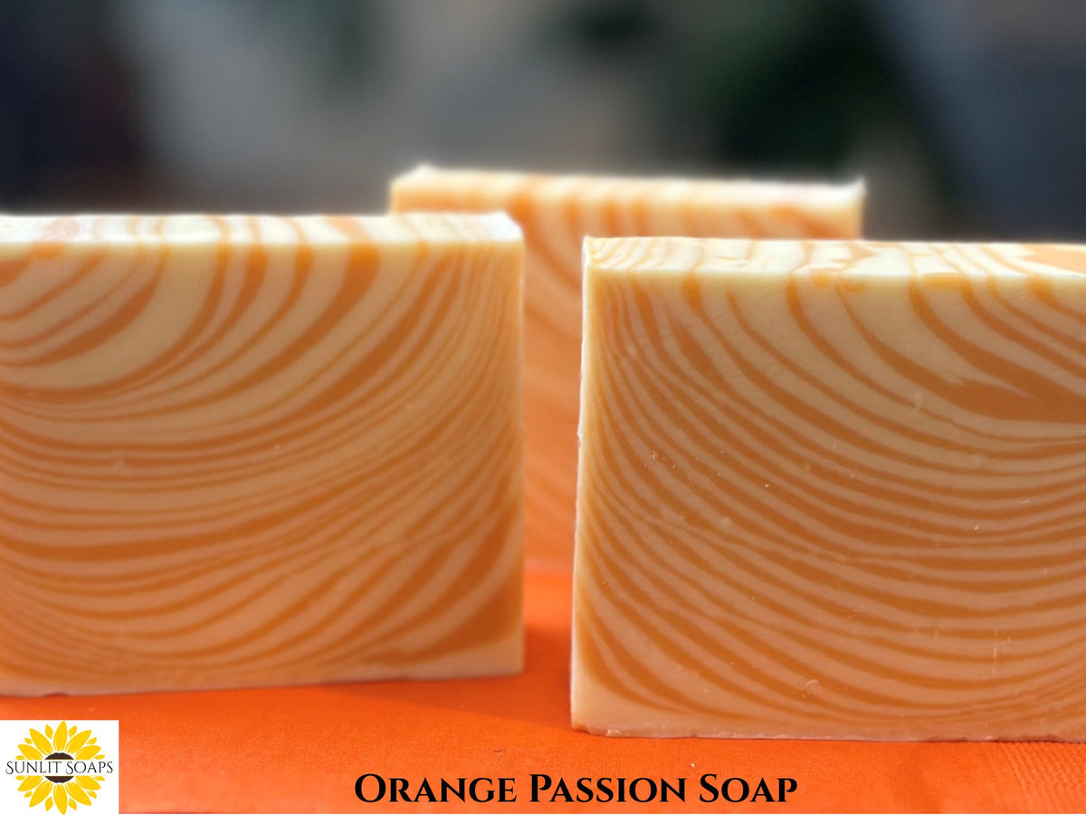 Three rectangular bars of orange passion soap with a wavy marbled pattern, displayed on an orange surface with a blurred background. A logo for 'Sunlit Soaps' is visible in the bottom-left corner, and the text 'ORANGE PASSION SOAP' is written at the bottom.