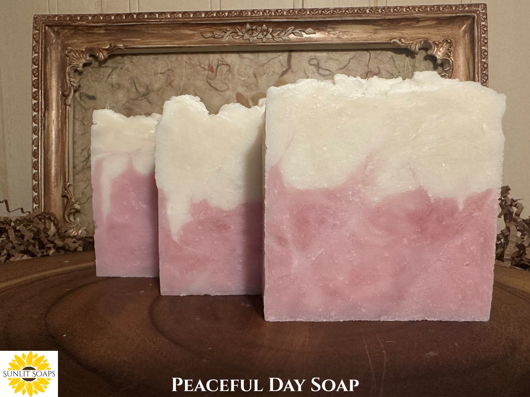 Three bars of pink and white soap on a wooden surface with an ornate wood frame in the background with Sunlit Soaps branding.