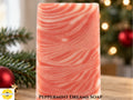 A rectangular bar of pink and white marbled soap standing upright on a wooden surface, with Christmas greenery and ornaments in the foreground and a softly blurred Christmas tree in the background. The words "Peppermint Dreams Soap" is at the bottom center, and the Sunlit Soaps logo is to the left.