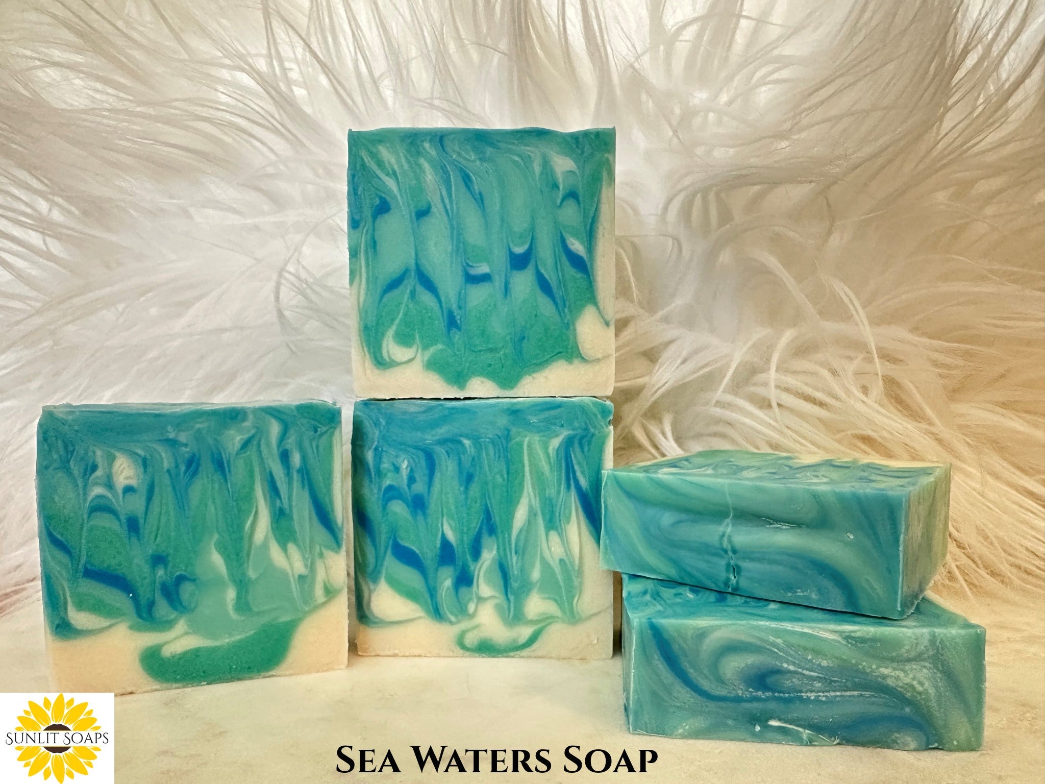Stack of blue and green swirled Sea Waters Soap bars with 'Sunlit Soaps' branding on a white fluffy background.