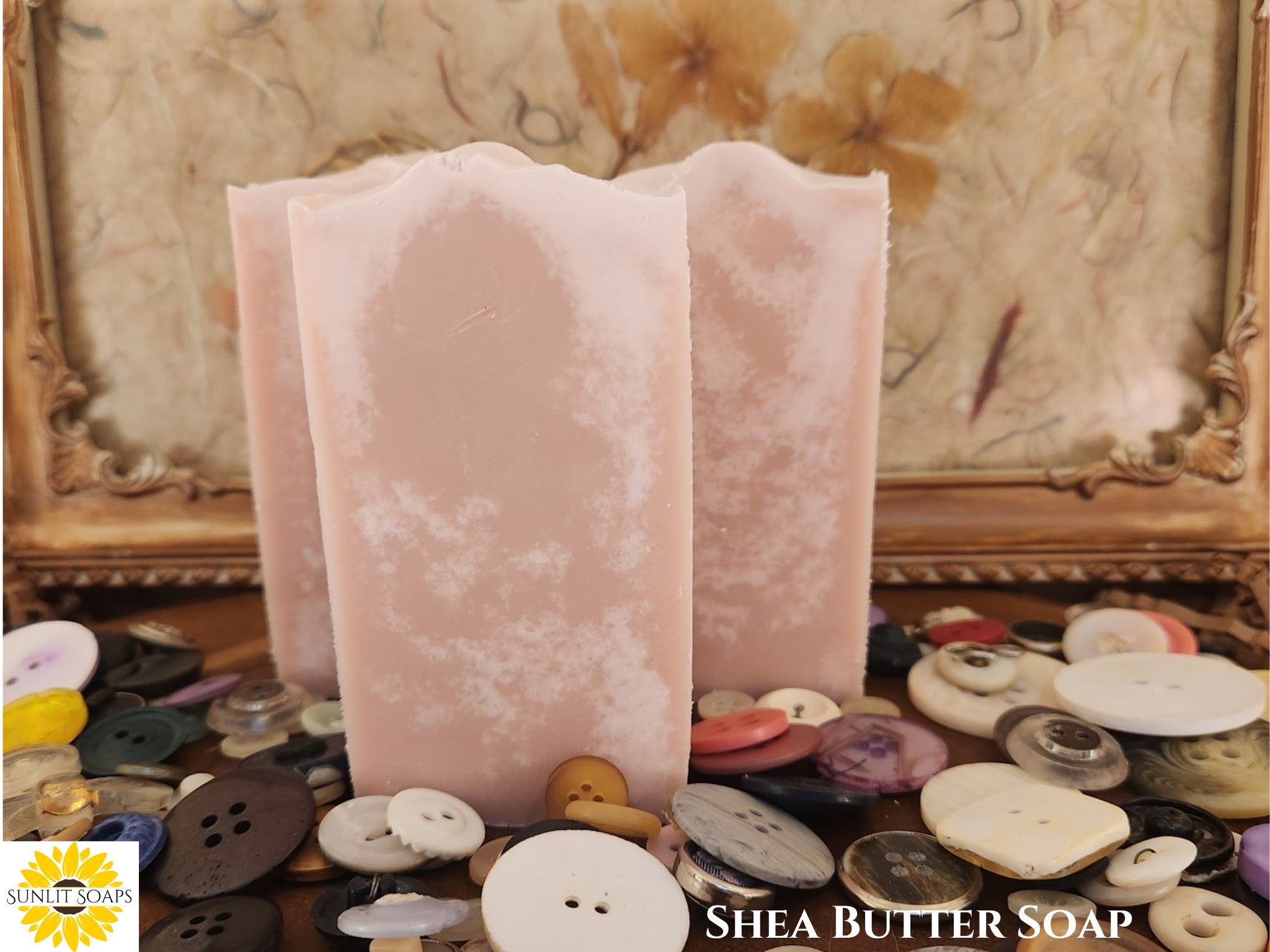 Pink tall and skinny Shea Butter soap bars with buttons scattered around in front of a decorative frame with Sunlit Soaps branding.