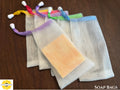 Several mesh soap bags with colorful strings on a wooden surface, featuring a logo and text for 'Sunlit Soaps'.