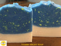 Two rectangular bars of handmade soap on a wooden surface. The front bar is fully visible, showing a cream-colored base and a marbled blue top layer with small yellow-green speckles.
The words "Starry Night Soap is on the bottom edge with the Sunlit Soaps logo in the left corner
